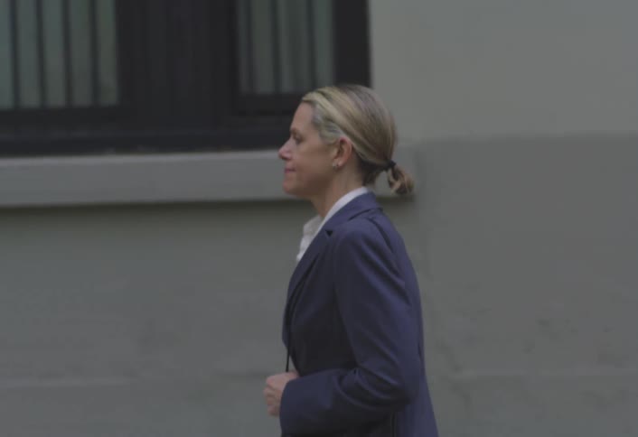 A woman in formal wear walks on the sidewalk past a large building