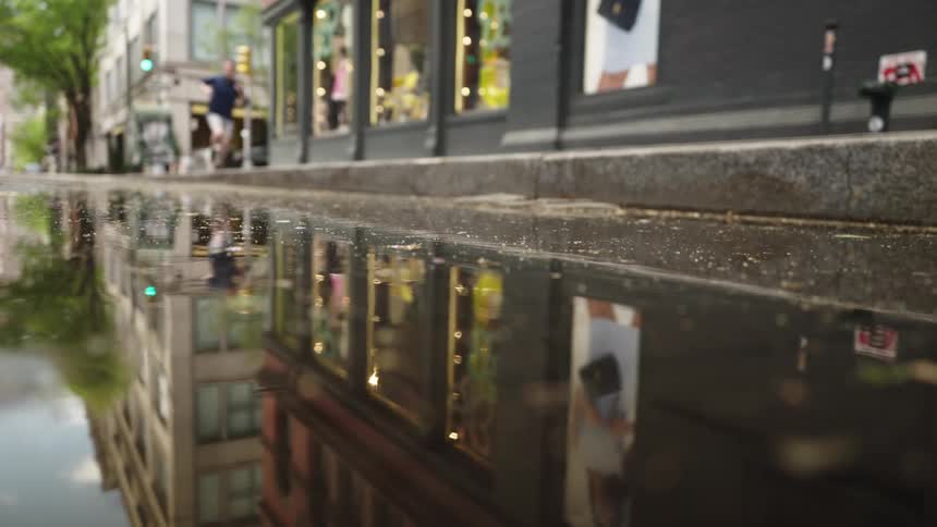 A city street and storefronts are reflected in a large puddle on the ground after rain