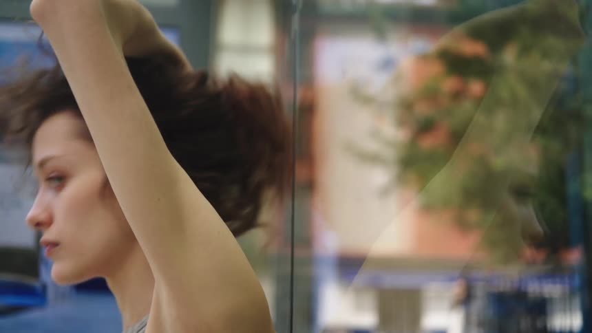 A woman with curly brown hair leans against a glass wall in the city her profile visible and a blurred reflection beside them