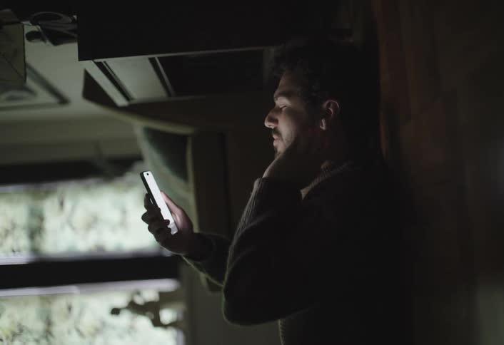 A man with curly hair and a beard sits in a dimly lit room illuminated by the bright screen of his smartphone as he scrolls through it with a thoughtful expression