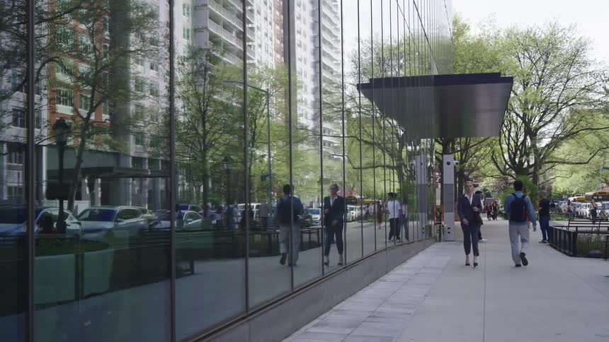 A woman in formal attire walks along a sidewalk next to a modern glass building