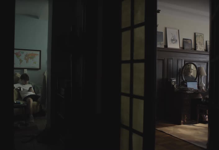 A darkened doorway frames two rooms with a person sitting on a chair looking at a phone in the left room and another person at a desk with a laptop in the right room