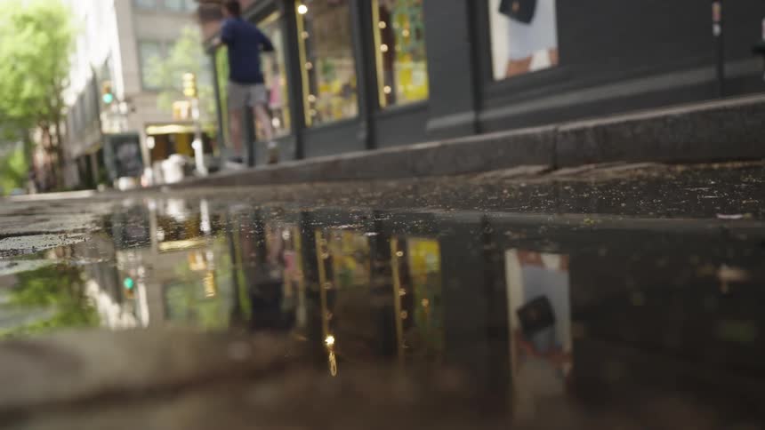 A shallow puddle on a city street reflects the brightly lit storefronts and the distant sidewalk as a person jogs by