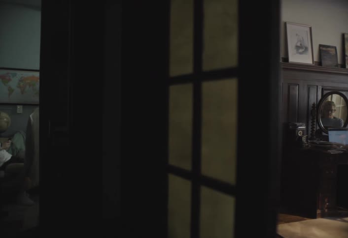 A person sits at a wooden desk with a laptop reflected on a mirror while in another dimly lit room a young man looks at a phone beneath a world map