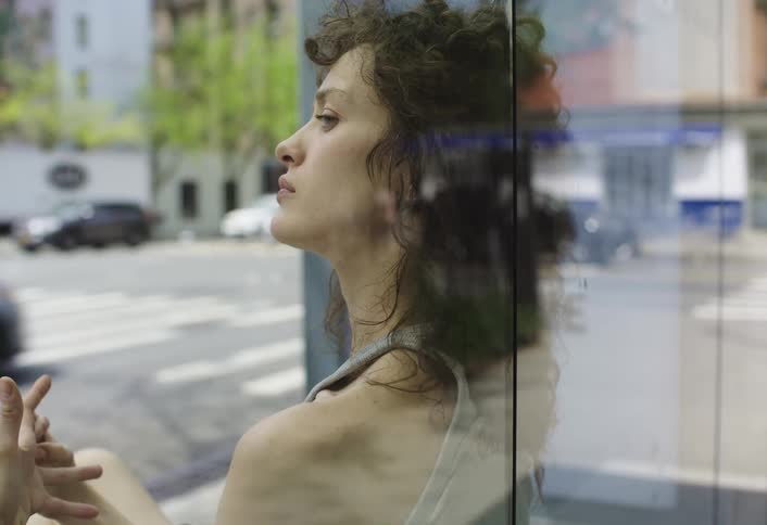 A woman with curly brown hair leans against a glass wall in the city
