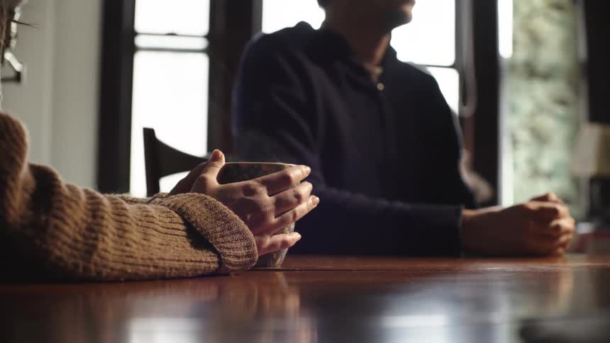 Two people sit at a wooden table one holding a mug and the other with hands clasped in a room with a window
