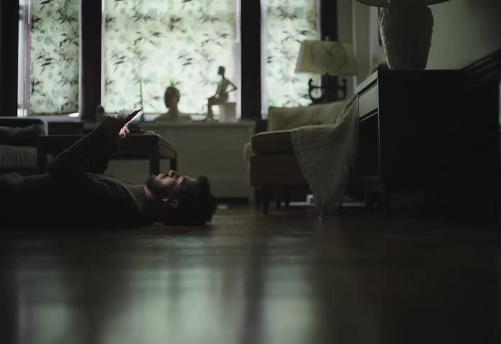 A person lies on a glossy wooden floor browsing a smartphone with furniture and a window featuring patterned curtains in the background