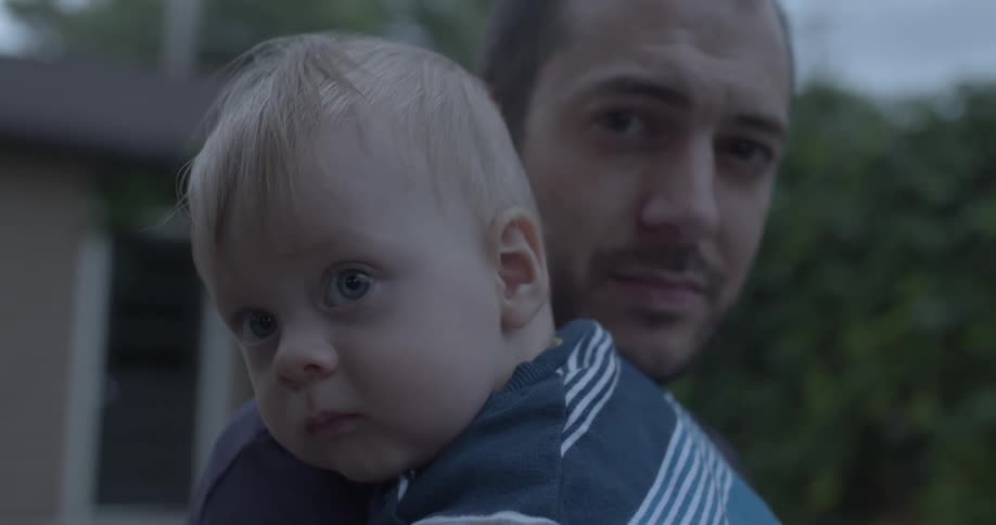 A blonde infant is held by an adult both looking directly forward with a blurred outdoor house in the background on a morning