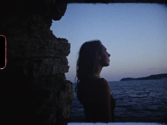 A young woman stands on the edge of a rock gazing at the ocean view in front of her at sunset