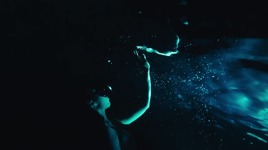 A woman is submerged in dark water illuminated by a vivid blue light that creates a shimmering trail of bubbles from the surface