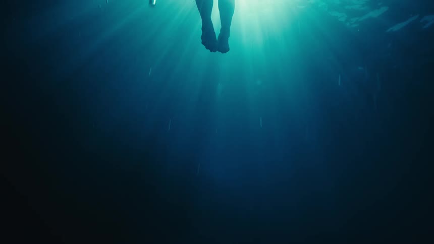A swimmers feet push off from a dark algaecovered rock silhouetted beneath a deep blue glow as sunlight pierces the water