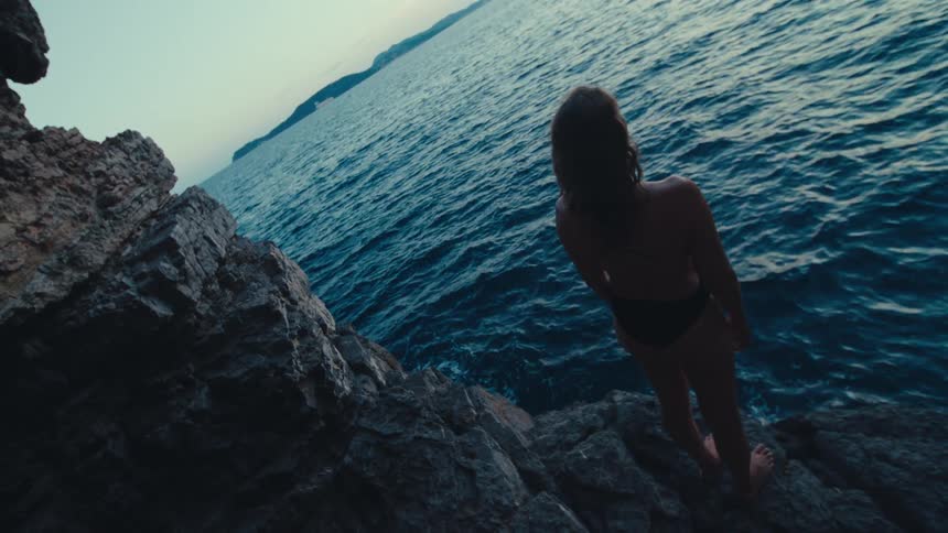 A woman stands on a rock beside the ocean at sunset