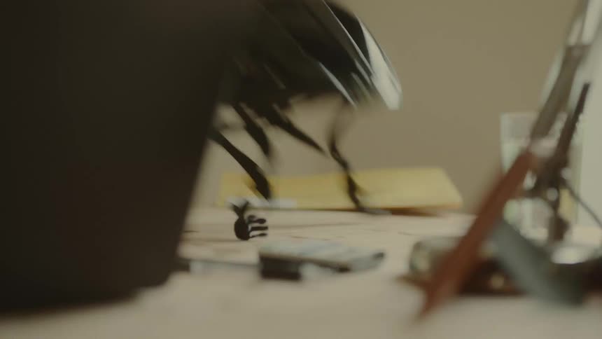 A hand picks up a black bicycle helmet from a desk that also holds other blurred objects and papers