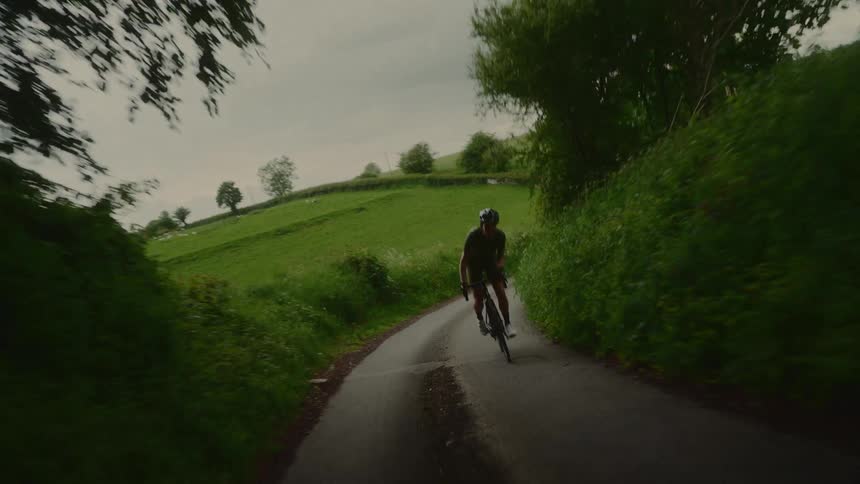 A person wearing a helmet cycles down a winding road surrounded by lush green hills and trees under a cloudy sky
