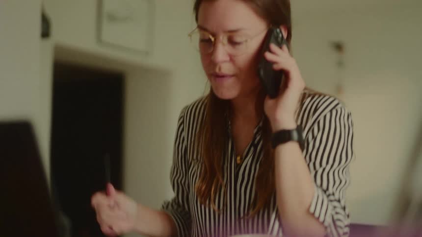 Woman sitting at a desk with an open laptop and on a call on her smartphone