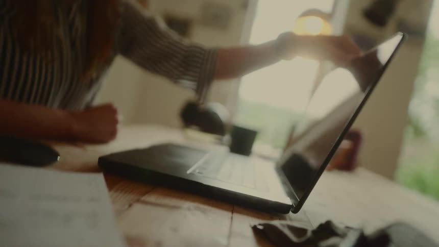 A persons hands are shown at a low angle working on a laptop with blurred background elements of a room and a bright window