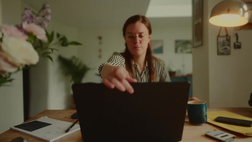 A woman with long brown hair and glasses opens a laptop on a wooden desk surrounded by office supplies and a vase of flowers in a warmly lit room
