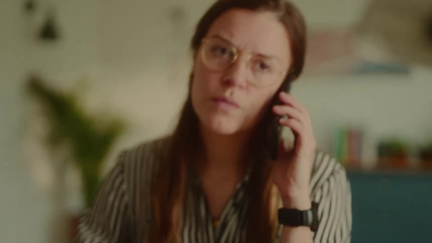 A woman wearing glasses sits behind a desk and picks up a phone to make a call