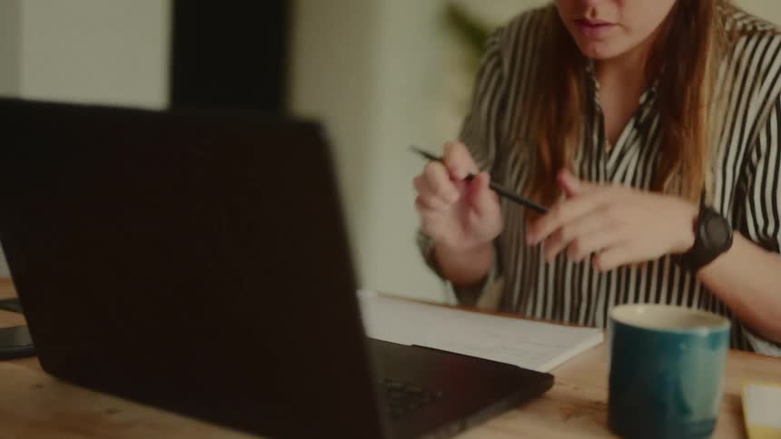 Woman holding a pen while sitting in front of an open laptop