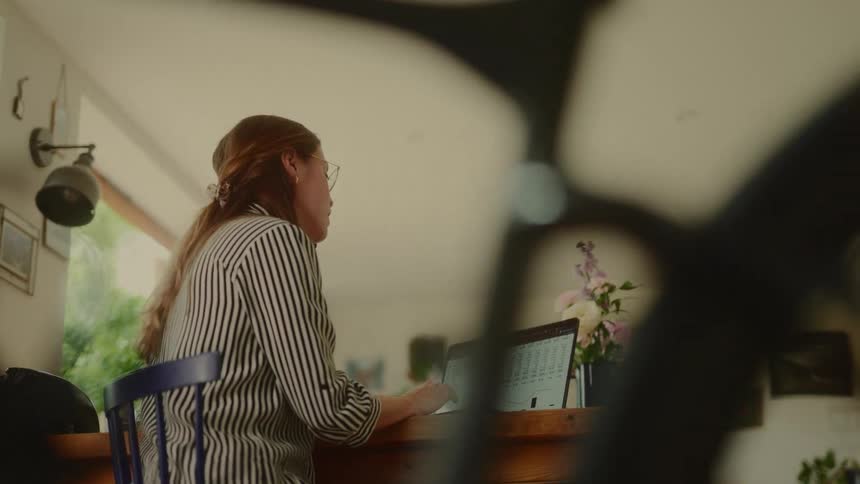 A woman with long hair wearing a striped shirt and glasses is seen from a low angle sitting at a wooden table with a laptop looking at the screen and briefly answering a phone call
