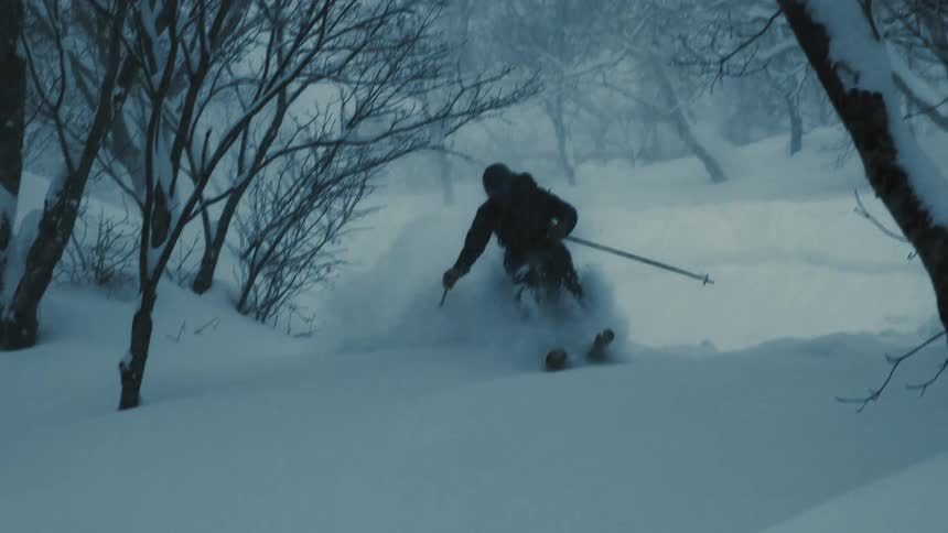 A lone skier descends a snowcovered slope through a sparsely treed area kicking up powder