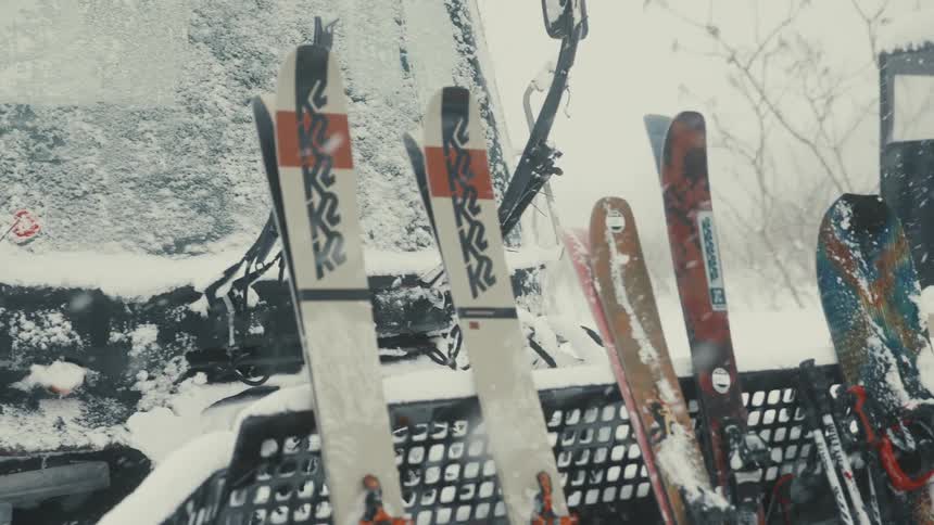 Skis and a snowboard covered in snow are propped against a vehicle or structure in a winter setting