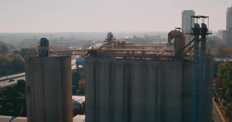 Large concrete silos with industrial structures on top stand prominently beside a highway with a cityscape and trees under a clear sky in the background