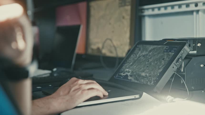 A hand operates a touchscreen device displaying satellite imagery or terrain maps