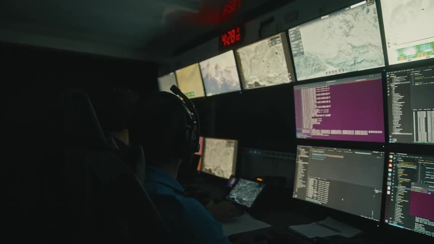 A person with headphones monitors multiple screens showing maps and data inside a control room