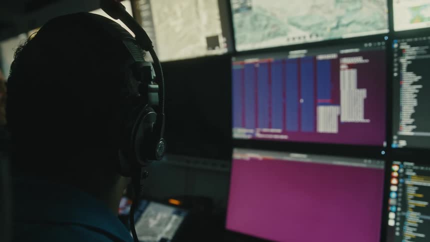 A person wearing a headset is seen from behind looking at a wall of multiple computer monitors displaying various data and maps in a dimly lit room