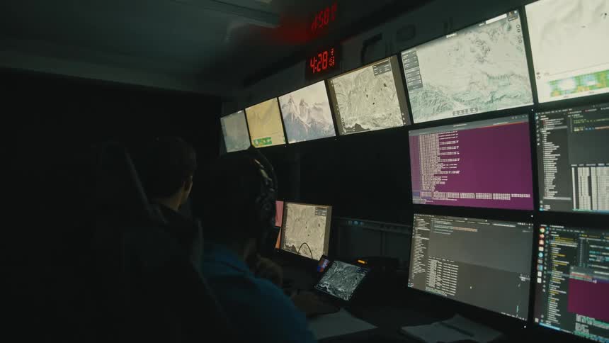 Two operators in a dark control room monitor multiple satellite and data screens