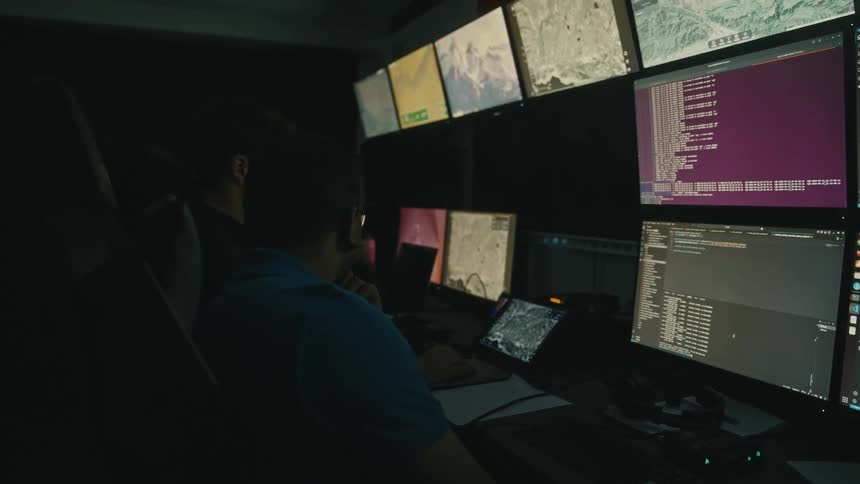 Coworkers observe one of the multiple computer monitors in a dimly lit computer room