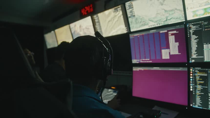 A person wearing a headset sits in front of a wall of multiple computer monitors displaying various data maps and information with another person visible in the background