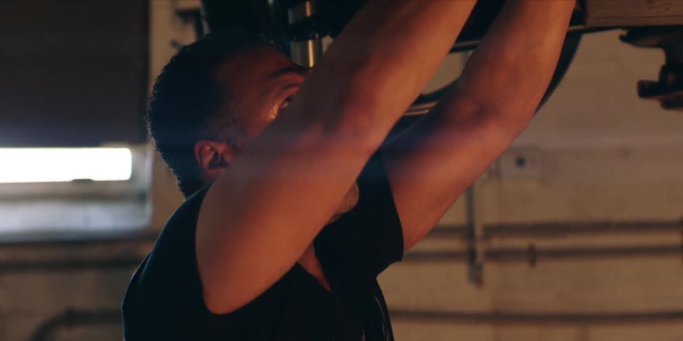 A man in a workshop fixing a car that is lifted above his head