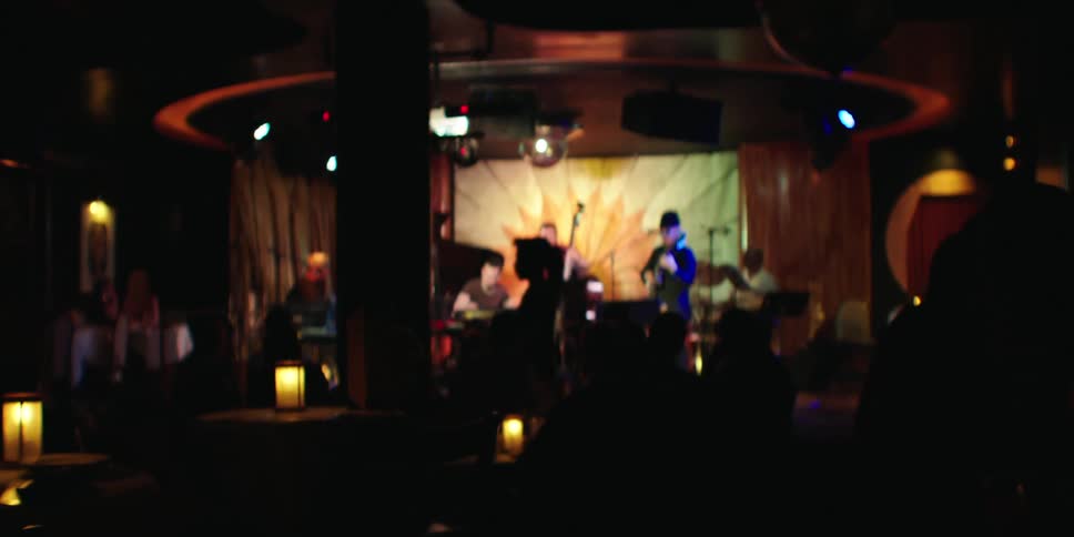 A blurred view of a band performing on a stage in a dimly lit venue with audience members in the foreground