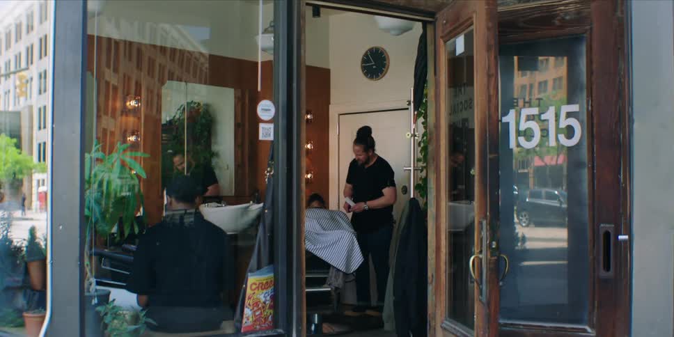 A man gets his hair cut by a barber in a barbershop at midday while other men sit patiently waiting for their turn