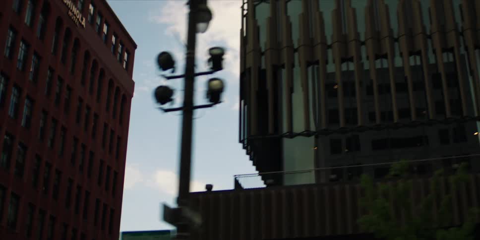 The scene pans right to reveal a modern glass building with a distinctive facade and reflections of the sky contrasting with a traditional brick building on the left under a partly cloudy sky