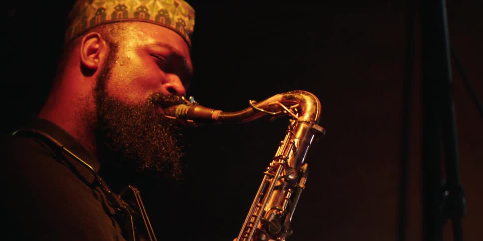 Black male musician plays a saxophone under a yellow stage light at night