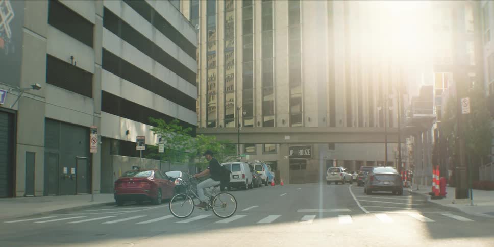 A man rides a bicycle through a sunlit city intersection surrounded by tall buildings