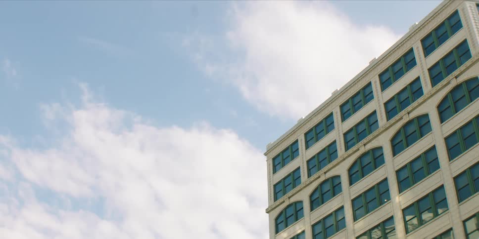 A multistory building with many windows is viewed from a low angle against a partly cloudy blue sky