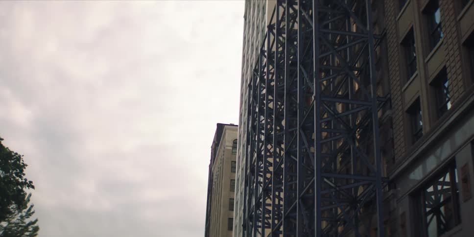 Metal framework attached to the front of a mid rise building towers over the city street on an overcast day