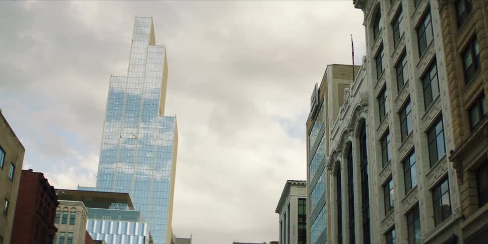 A modern glass skyscraper with reflections dominates the left side of the frame under a cloudy sky while a more ornate traditional building with an american flag fills the right side