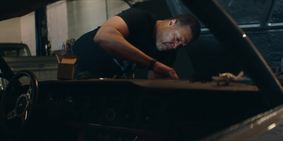 A mechanic with a focused expression leans into the passenger side of a classic car to work on the interior inside a dimly lit garage