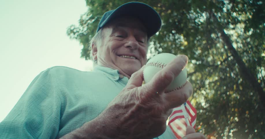 An older man wearing a baseball cap and light blue shirt smiles broadly while holding a baseball and a small american flag looking up towards a bright leafy sky