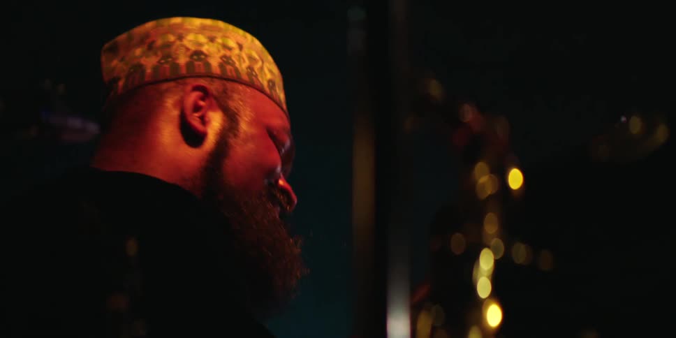 A man with a full beard and a patterned cap is bathed in warm reddish light from the left casting most of his face in shadow as he looks towards the right where blurry golden lights create bokeh against a dark background