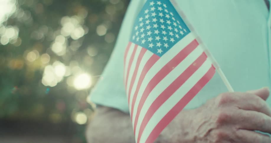 An older man holds an american flag then reveals a broad smile as the camera tilts up with a bright lens flare illuminating the blurred green background