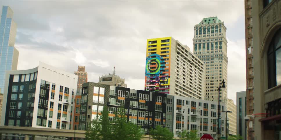 Street leads to a high rise with a colorful facade that towers over several of the nearby downtown buildings on an overcast day