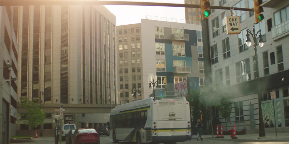 A bus driving along a city road at midday while a group of people ride scooters on the roadside with the traffic light ahead glowing green