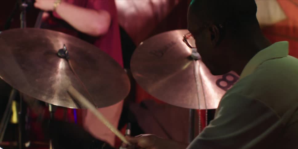 A black drummer passionately plays the drums during a concert performance while another man beside him plays the guitar