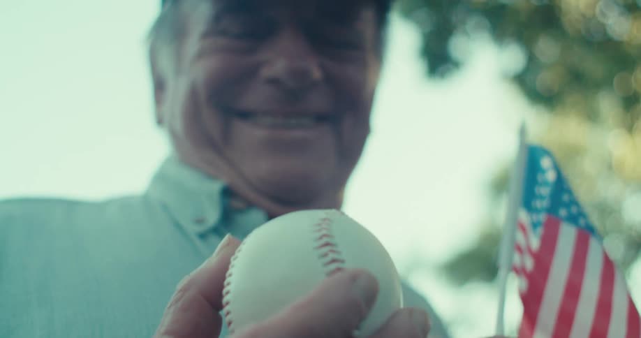An elderly man smiling as he looks at a baseball in his right hand holding a small flag on his left arm while outdoors at midday
